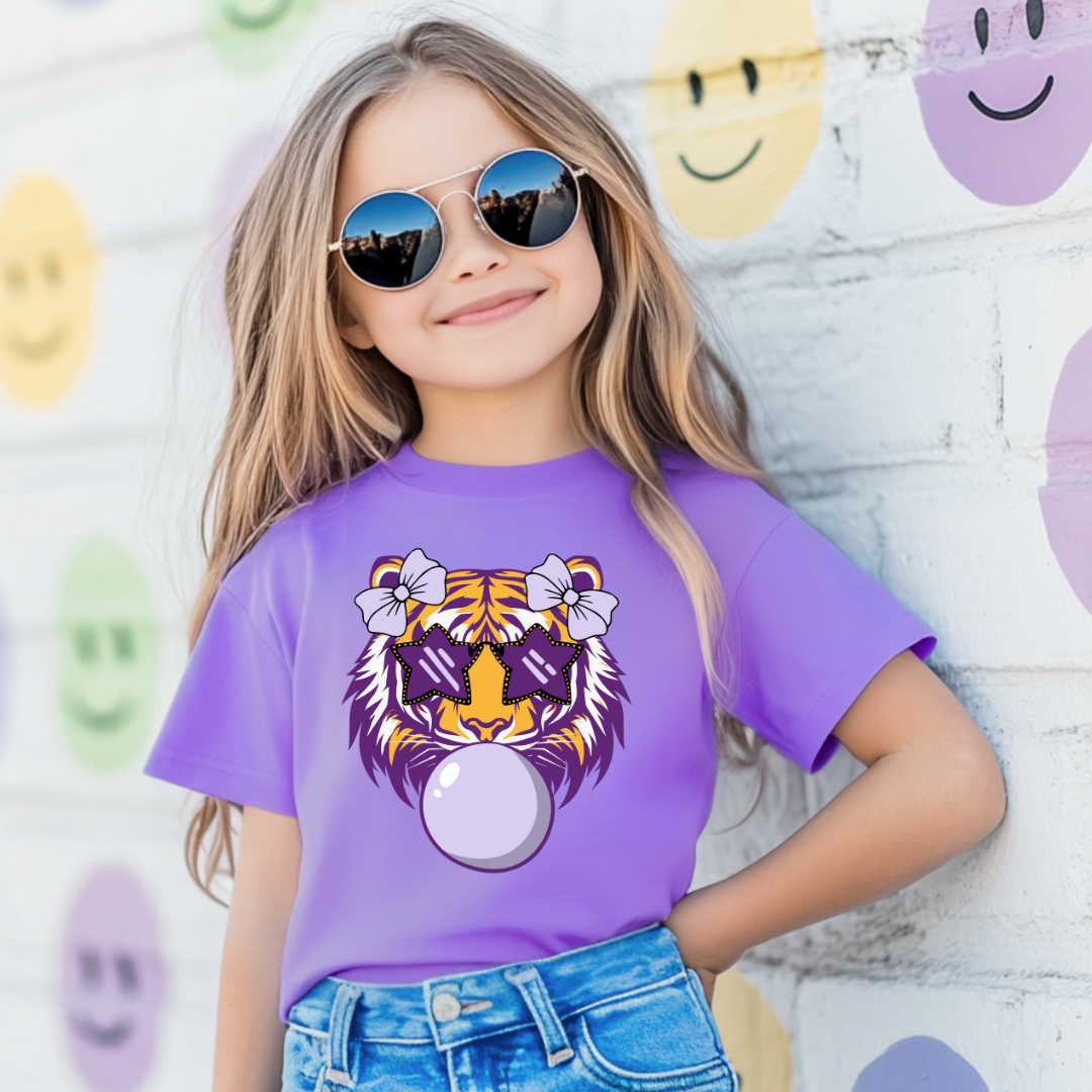 Child wearing a purple t-shirt with a tiger design, standing against a wall with smiley face decorations.
