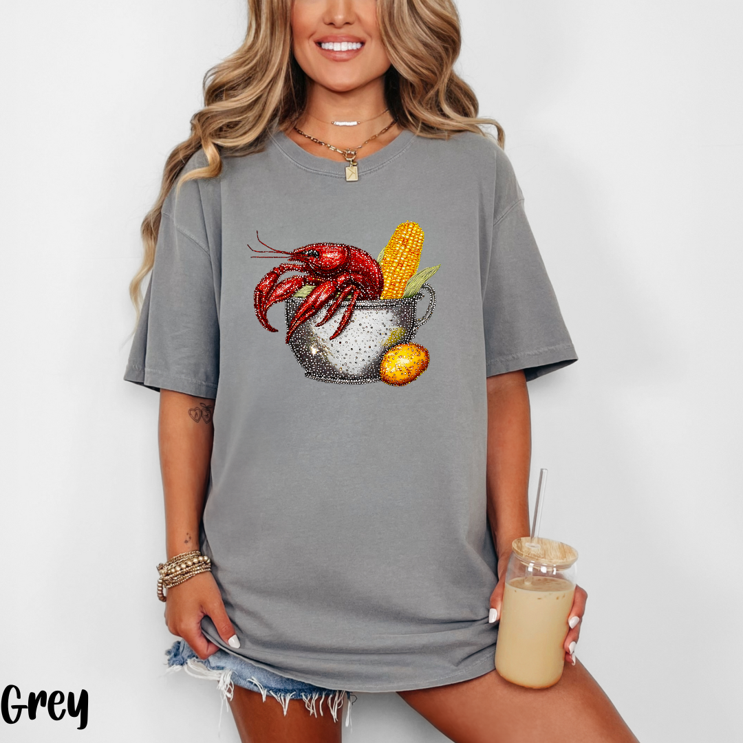 Woman wearing a gray t-shirt with a crawfish and corn graphic, holding a drink.