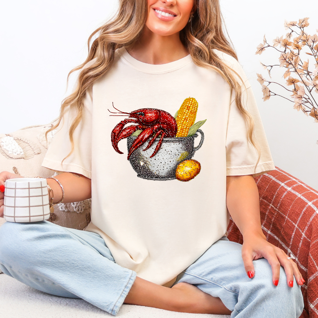 Woman wearing a white t-shirt with a crawfish and corn graphic, holding a mug.
