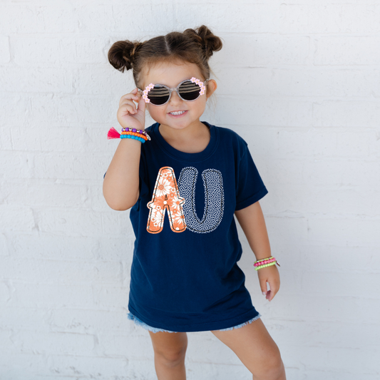 Child wearing a navy blue t-shirt with 'AU' design, holding sunglasses, against a white brick wall.