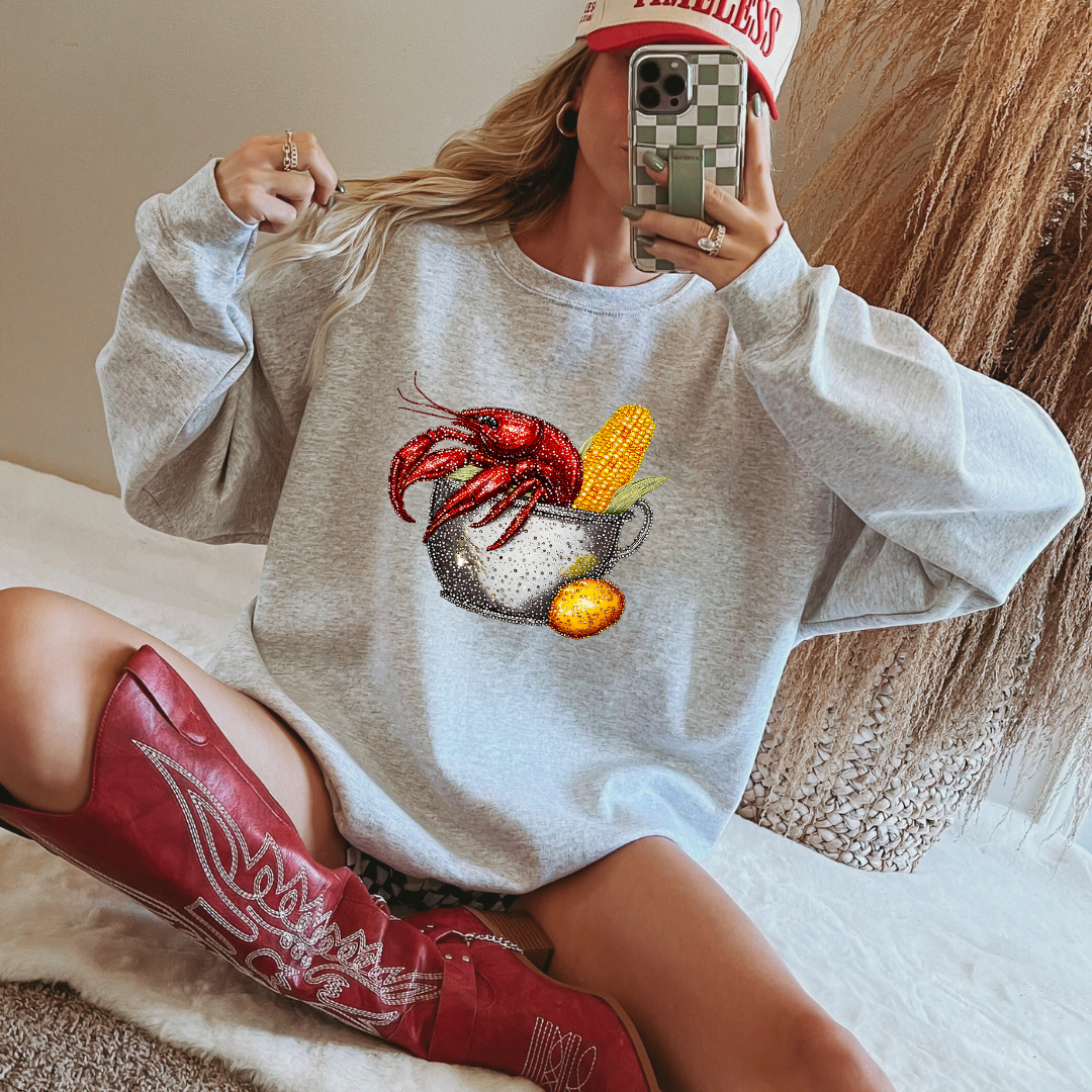Person wearing a sweatshirt with a crawfish and corn graphic, sitting on a bed.