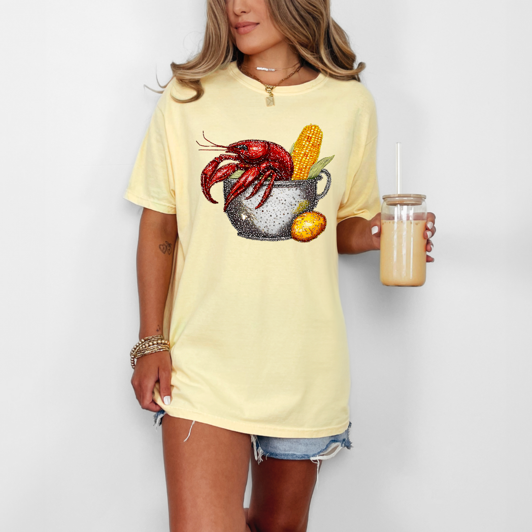 Person wearing a yellow t-shirt with a crawfish and corn design, holding a smoothie.